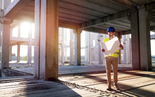 Baret ve güvenlik yeleği giyen inşaat mühendisi, çok katlı kaba beton bir binada gün batımında şantiye planlarını inceliyor